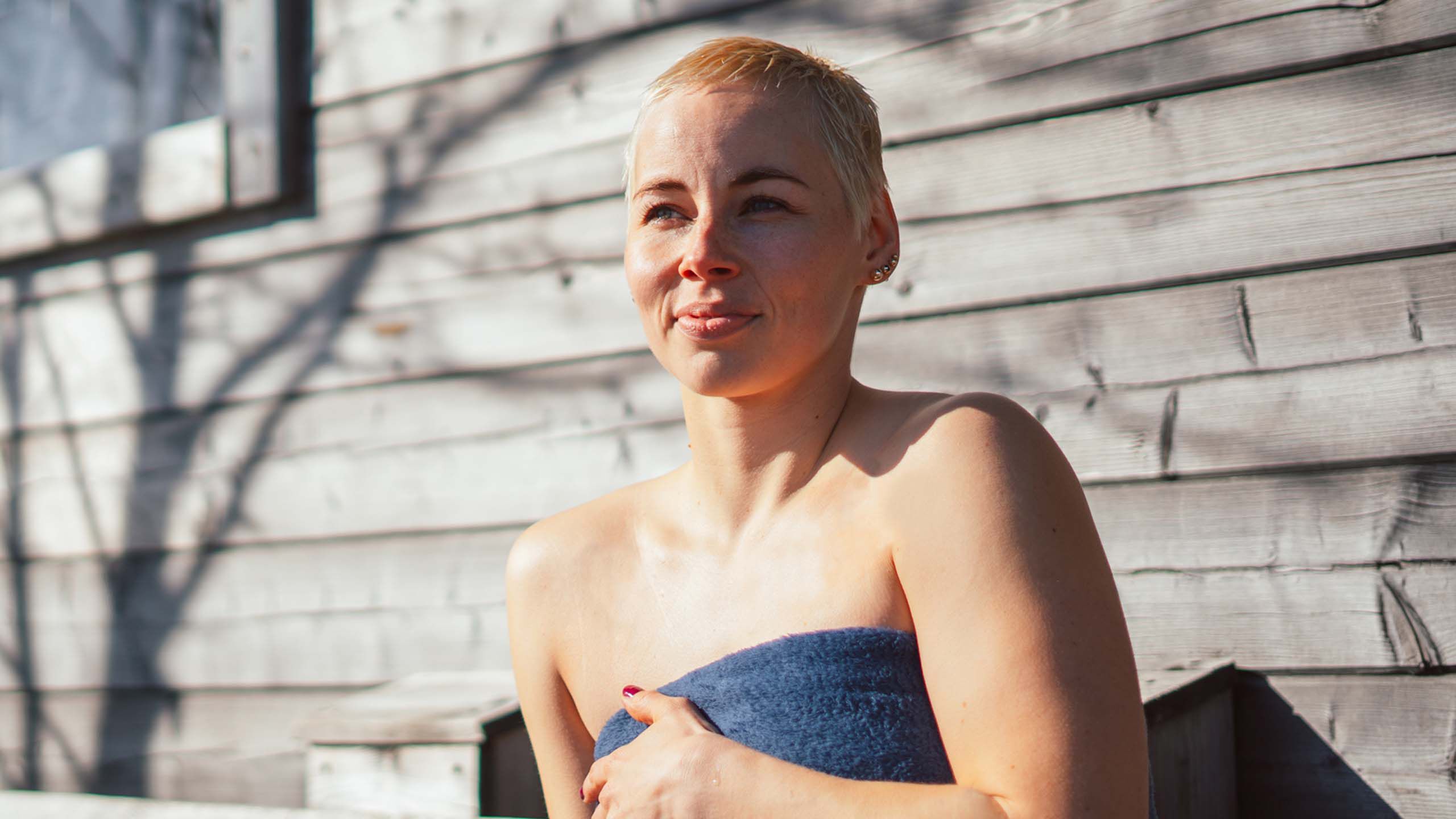 Nainen istuu saunan edess&auml;, pyyhe p&auml;&auml;ll&auml;.